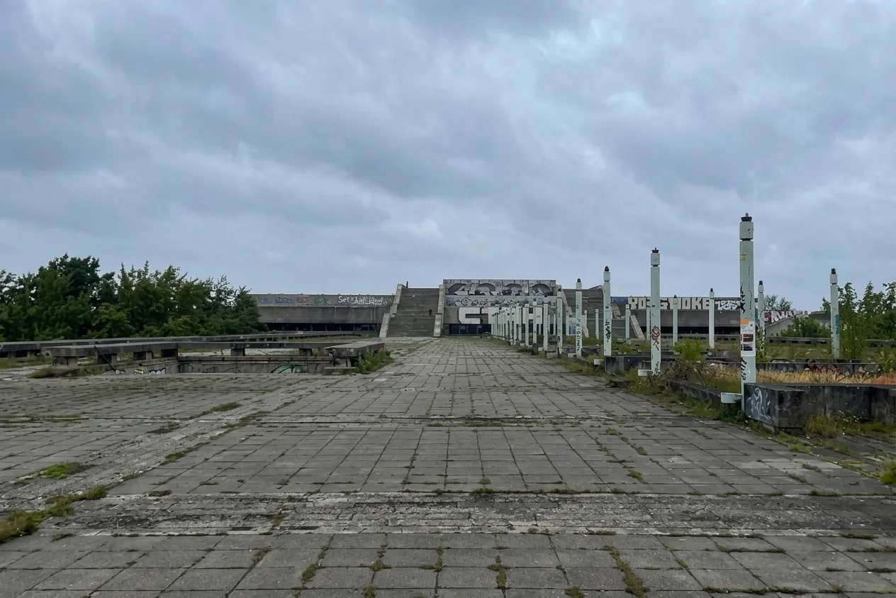 Weite, von Unkraut durchzogene Betonfläche unter grauem Himmel, rechts eine Reihe Laternenmasten, am Ende Treppen und Wände voller Graffiti.